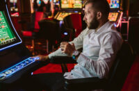 A man in a white shirt drinks his whiskey and plays a slot machine